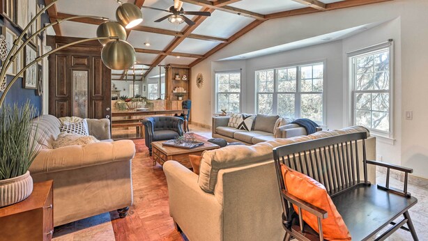 Ein geräumiges Wohnzimmer mit hohen Decken, Holzbalken und großen Fenstern. Mehrere Sitzbereiche mit Sofas und Sesseln, einem Couchtisch und einem Bücherregal.