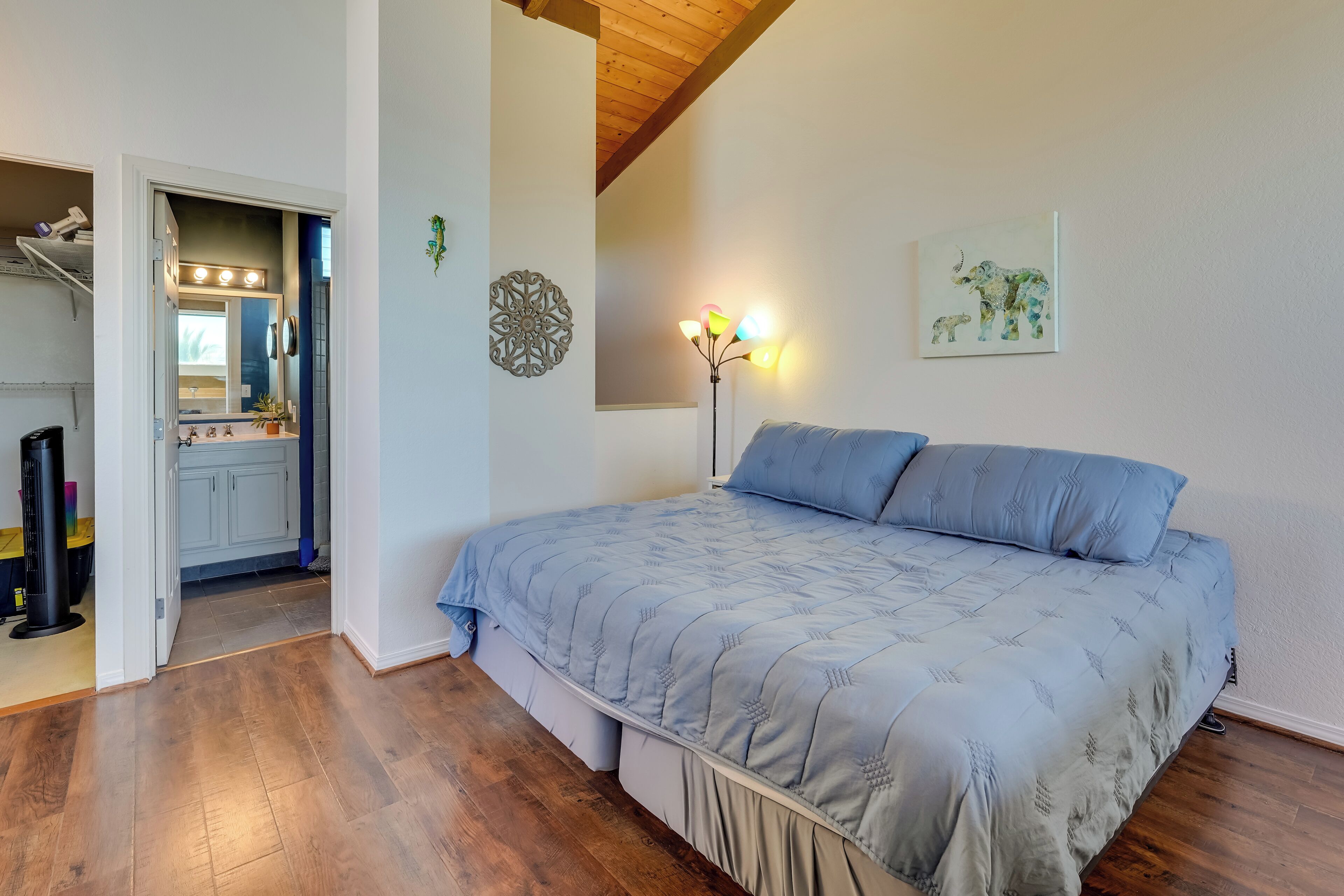 A bedroom with a bed, wooden flooring, a ceiling fan, a wall-mounted light, and a door leading to a bathroom.
