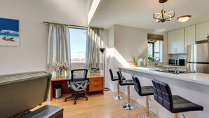 A modern kitchen with white cabinets, a stainless steel refrigerator, and a microwave. There is a wooden desk with a chair and a small trash can. A large window with curtains allows natural light into the room. - Top-floor Kailua Bay Resort Condo w/ Ocean Views! (Kailua-Kona)