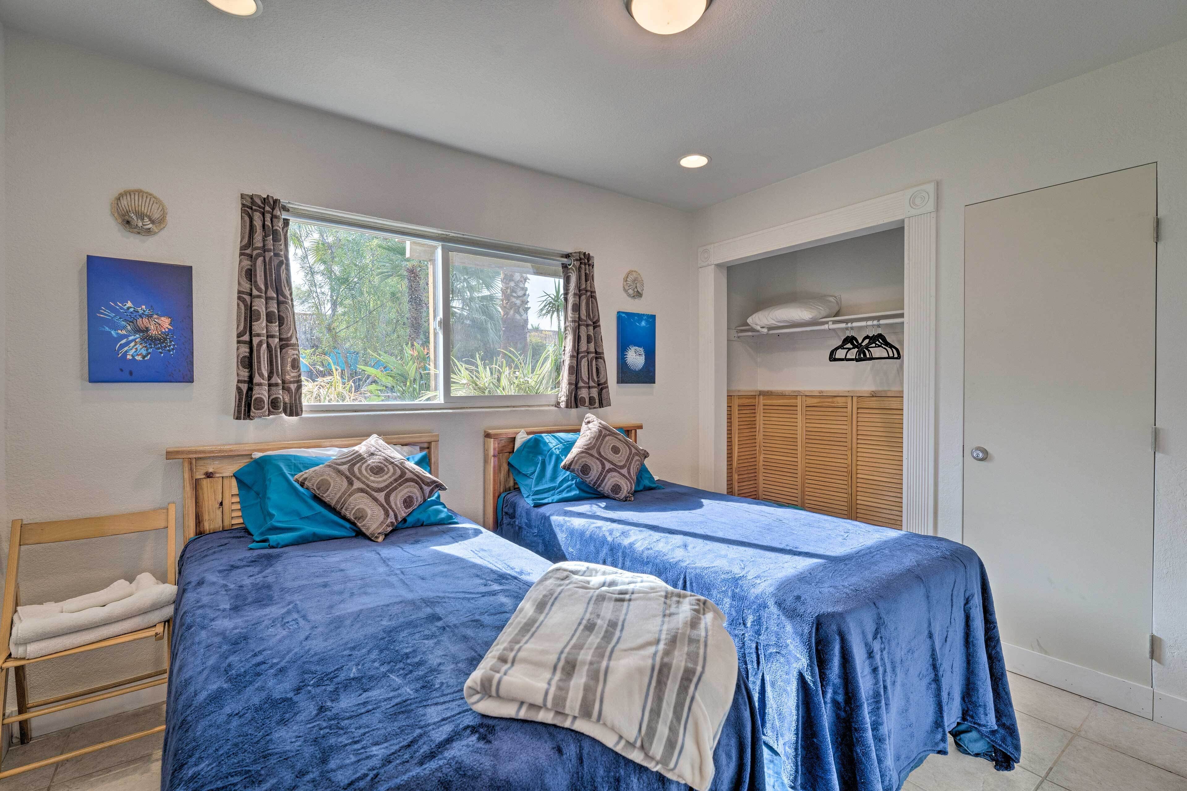 A room with two beds, a window with curtains, a wooden wardrobe, and a shelf with hanging items.