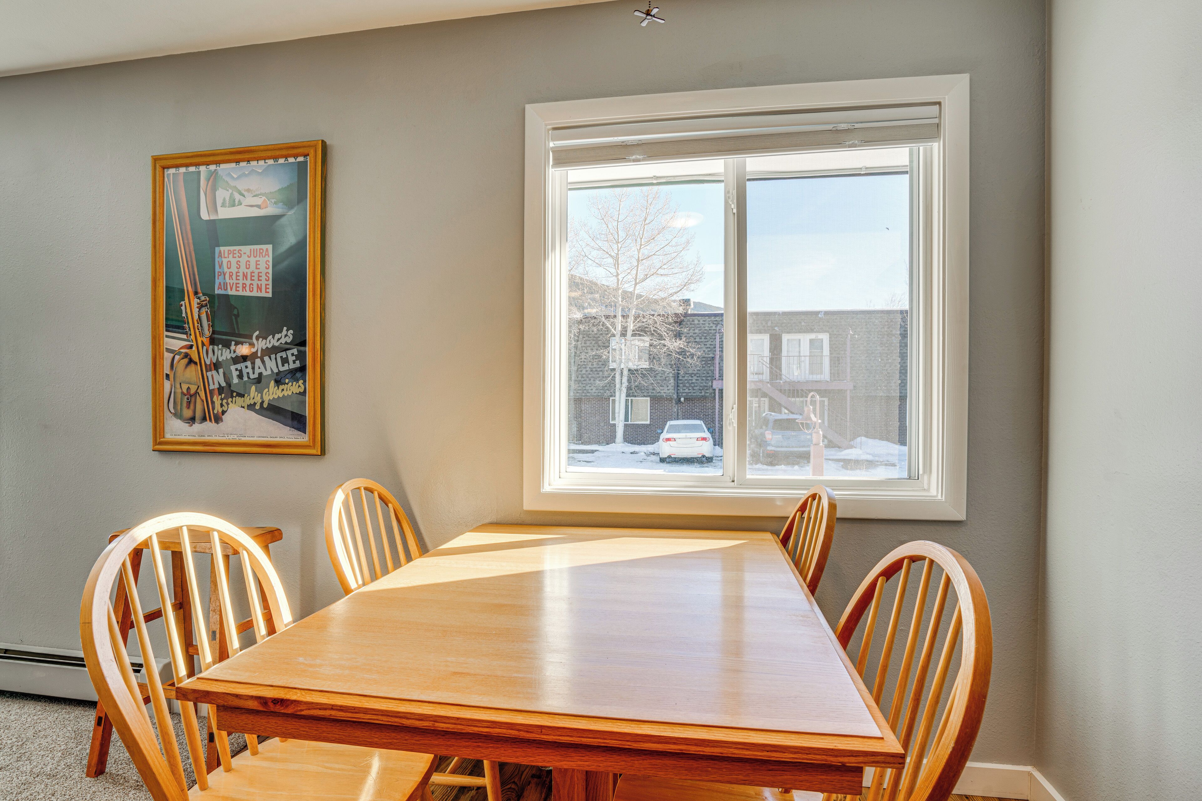 Een eethoek met een houten tafel en stoelen, een raam met uitzicht op een besneeuwde straat en een lijst met een poster aan de muur.