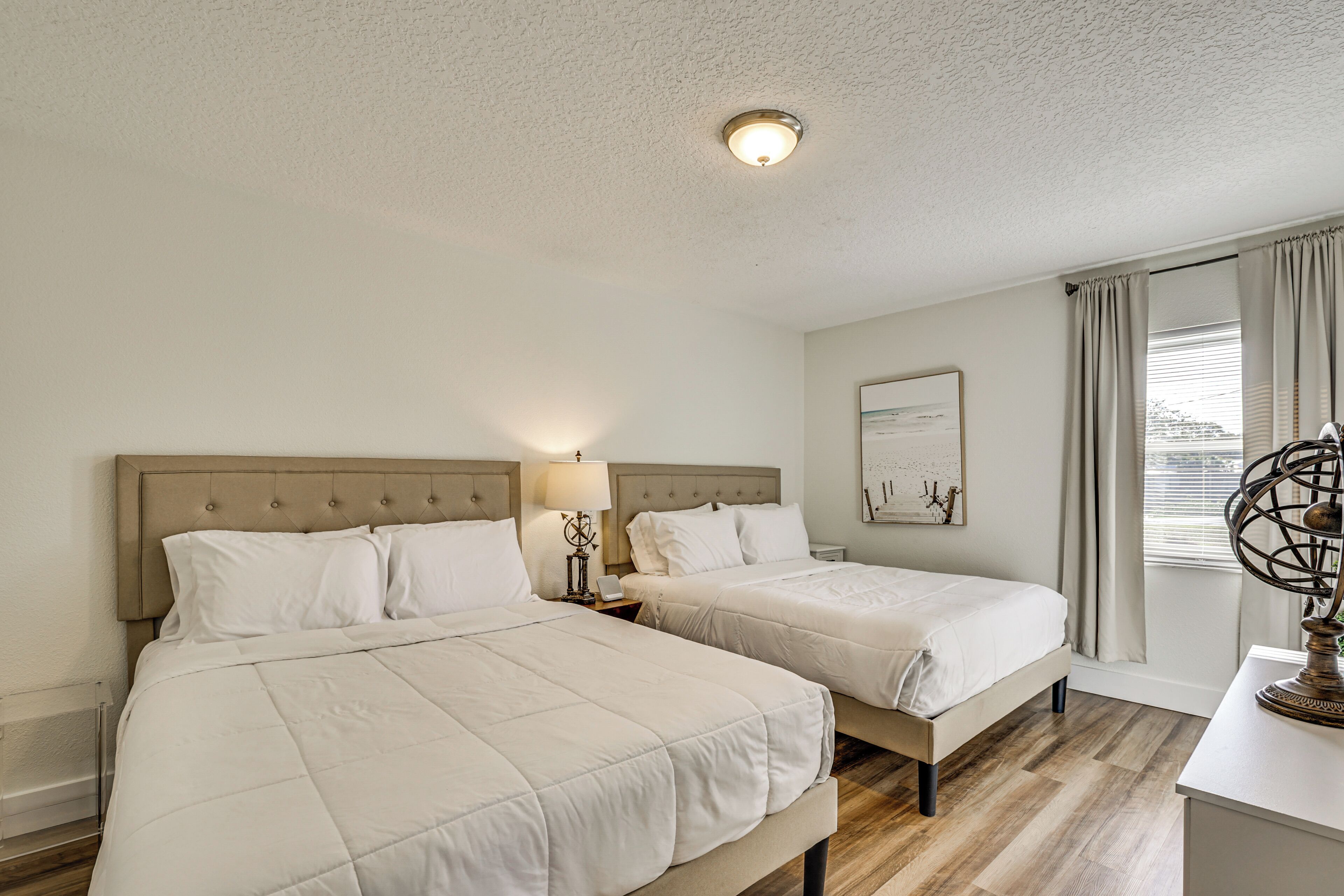 A hotel room with two beds, a ceiling light, a window with curtains, and a wall-mounted artwork.