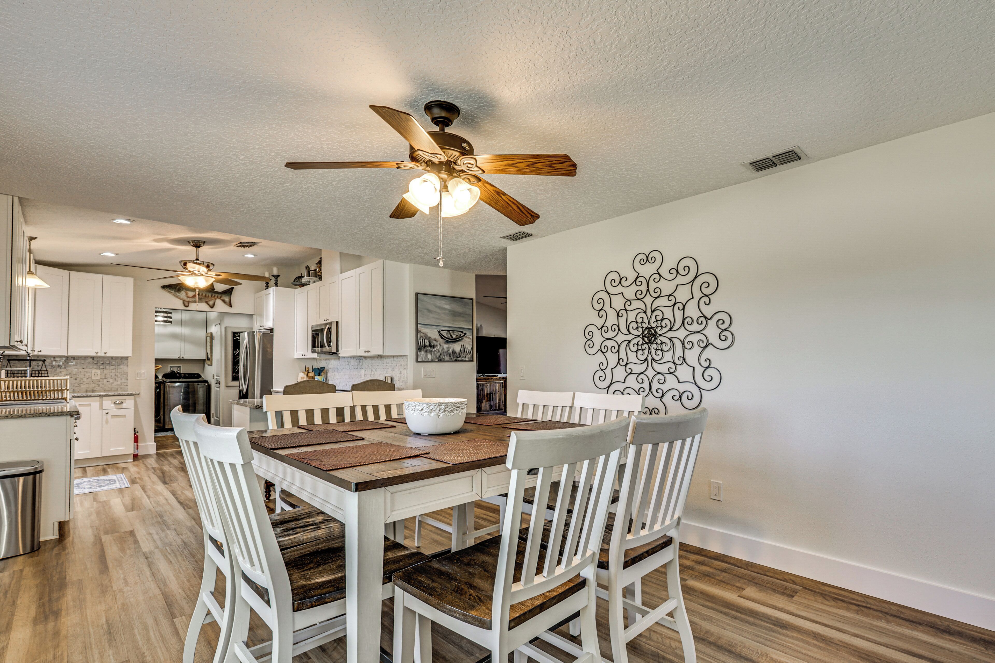 A modern kitchen with white cabinets, a wooden dining table, and a ceiling fan.