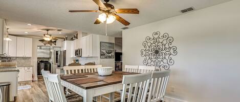 A modern kitchen with white cabinets, a wooden dining table, and a ceiling fan.