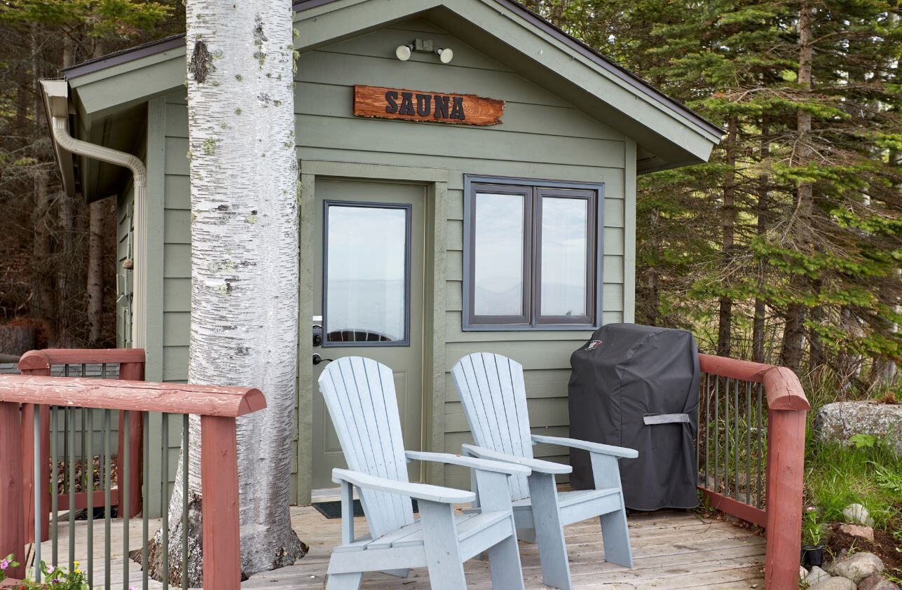 A small cabin with a sign reading "SAUNA" above the door, two Adirondack chairs on the deck, and a covered grill.