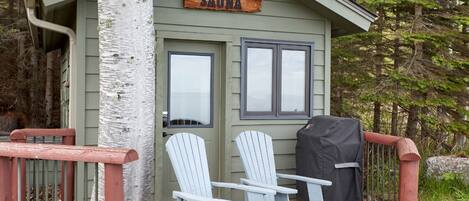 A small cabin with a sign reading "SAUNA" above the door, two Adirondack chairs on the deck, and a covered grill.