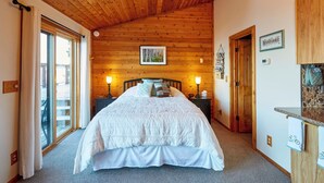 Een slaapkamer met een houten plafond, een groot bed met wit beddengoed, twee bedlampen, een ingelijst schilderij aan de muur, en een houten deur die naar een andere kamer leidt.