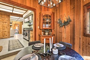 Interior de madera con una mesa de comedor preparada para cuatro, una cocina al fondo y una lámpara en el techo.