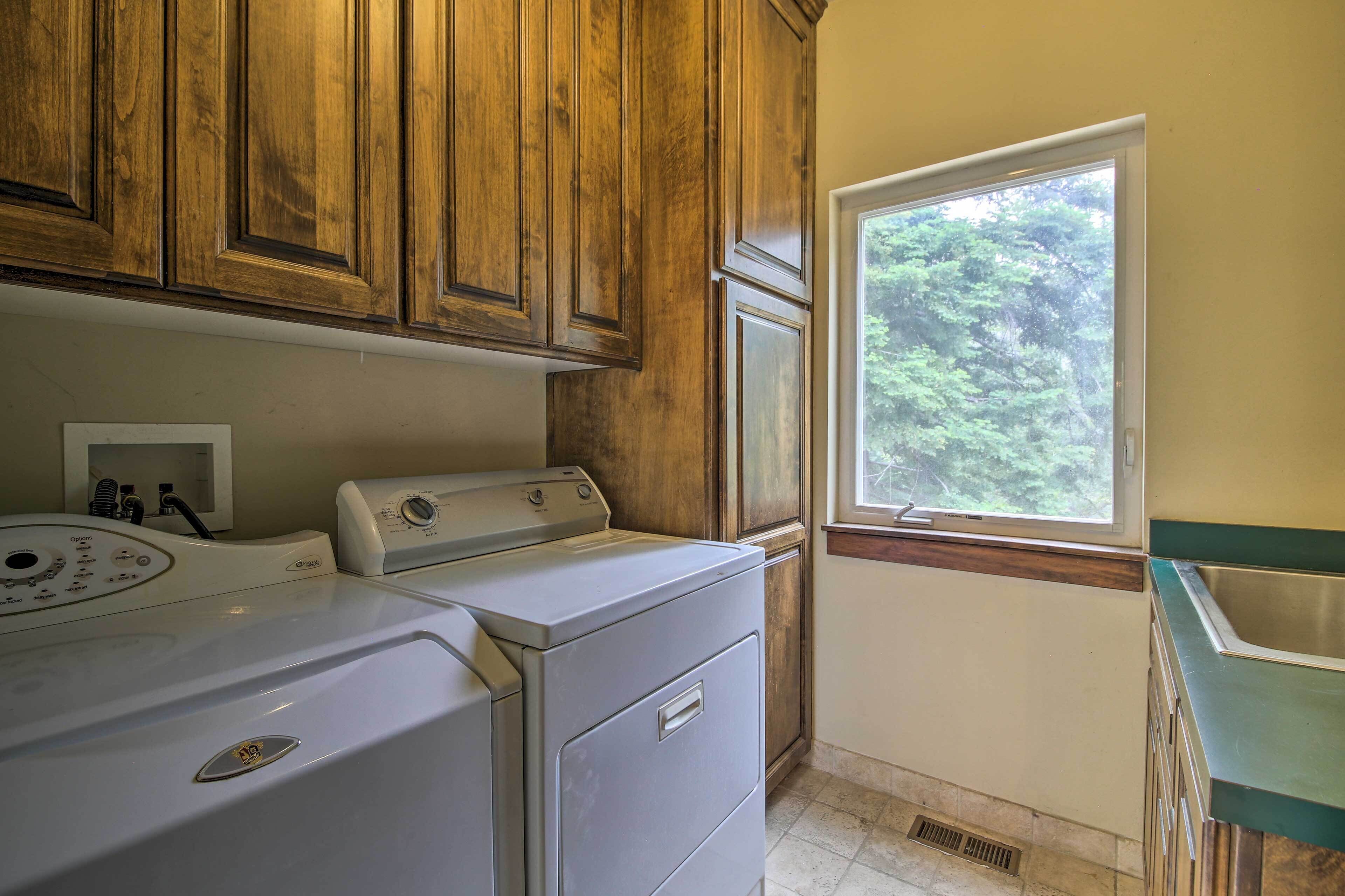 A laundry room with wooden cabinets, a washing machine, a dryer, and a window with a view of trees.