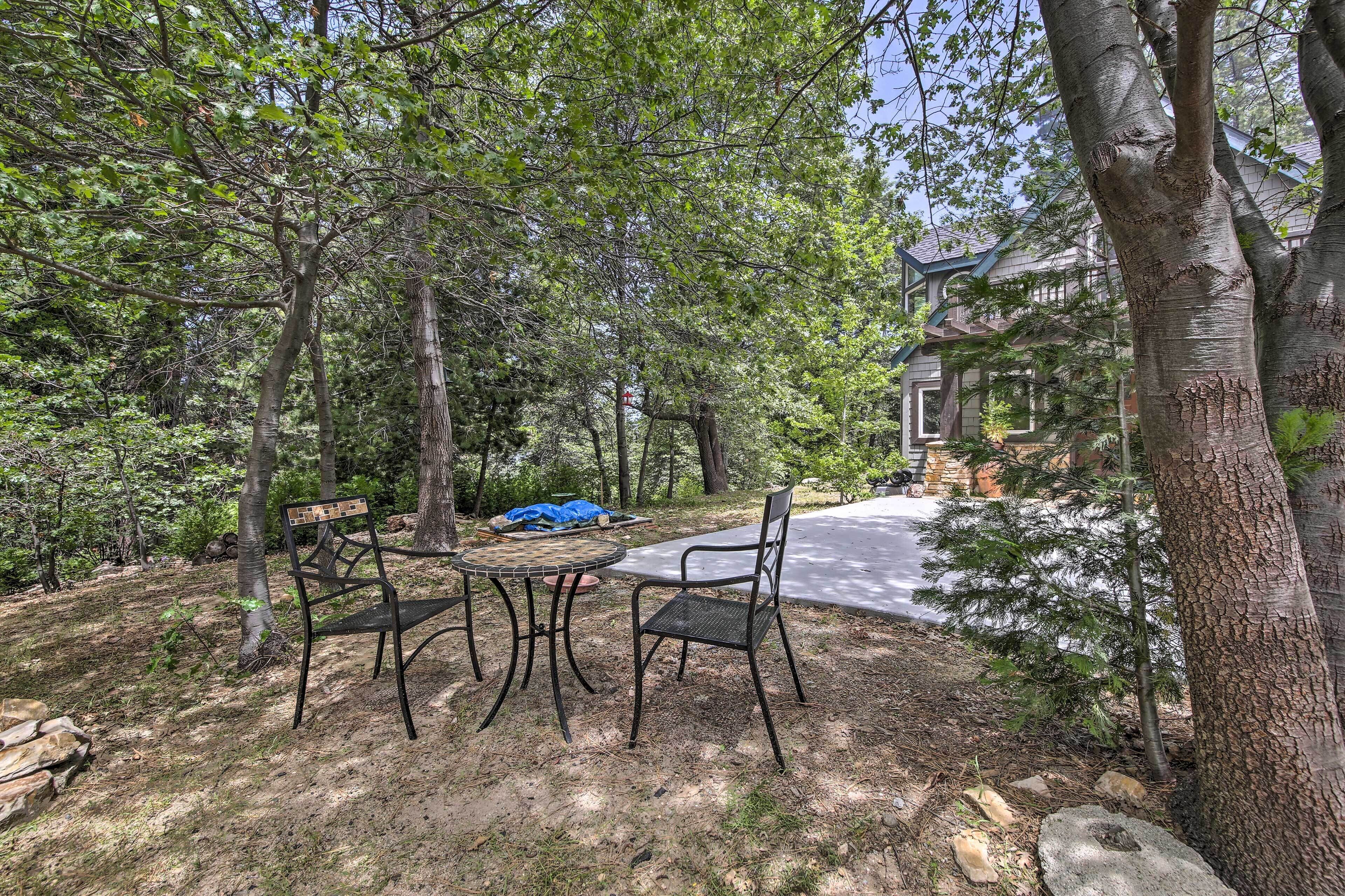 Une terrasse avec des chaises et une table en métal, entourée d’arbres et avec une maison en arrière-plan.