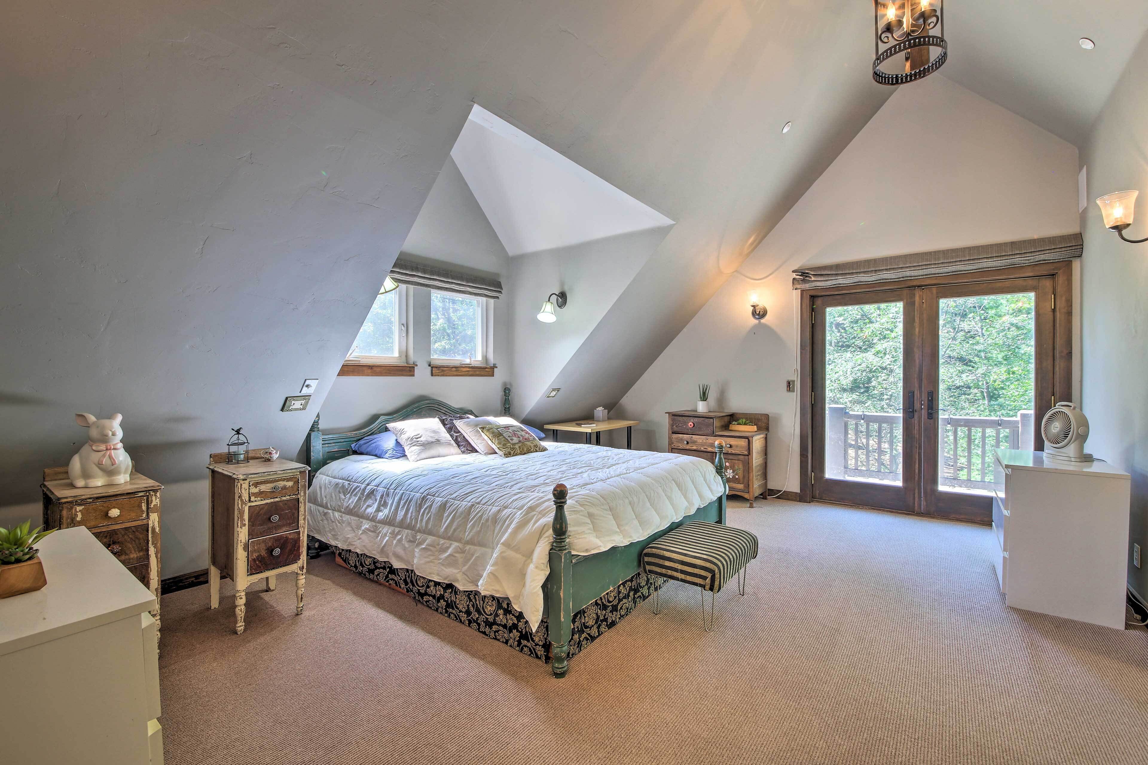A bedroom with a sloped ceiling, a large bed with a green bedspread, two bedside tables with lamps, a bench, and a view of the outdoors through a large window.