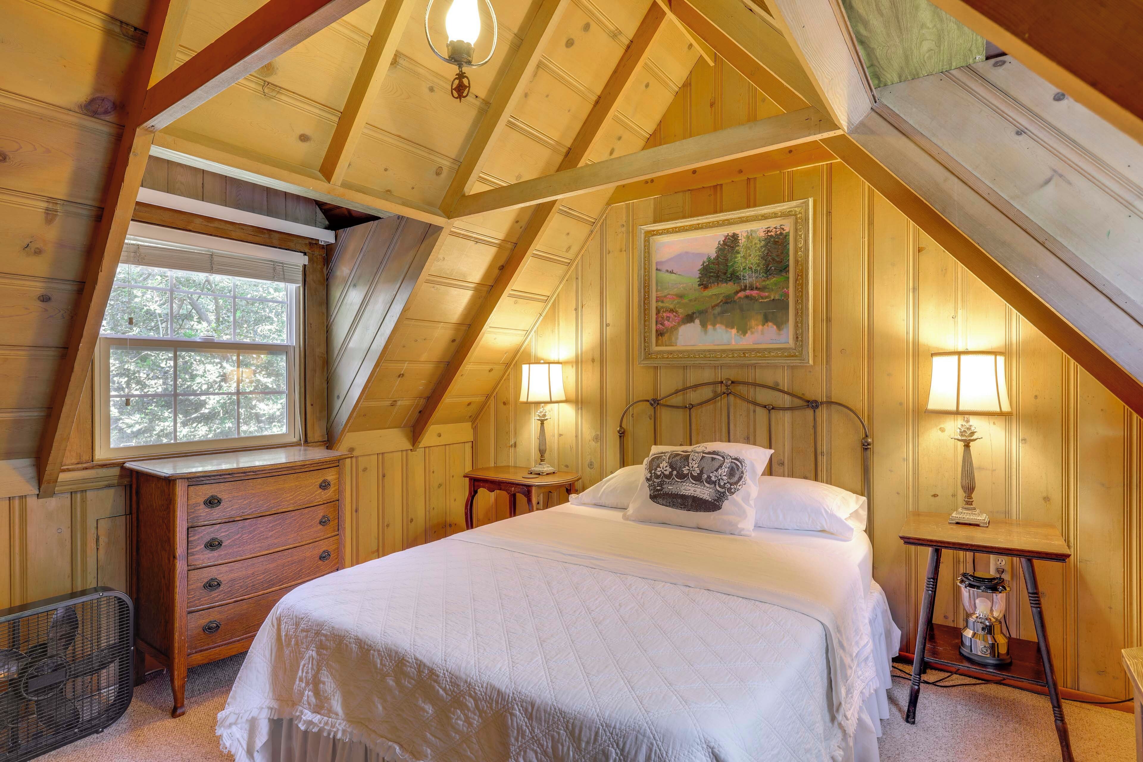 Une chambre confortable située dans les combles, avec un plafond en bois, un grand lit recouvert de linge de lit blanc, deux lampes de chevet et une fenêtre donnant sur la vue des arbres.