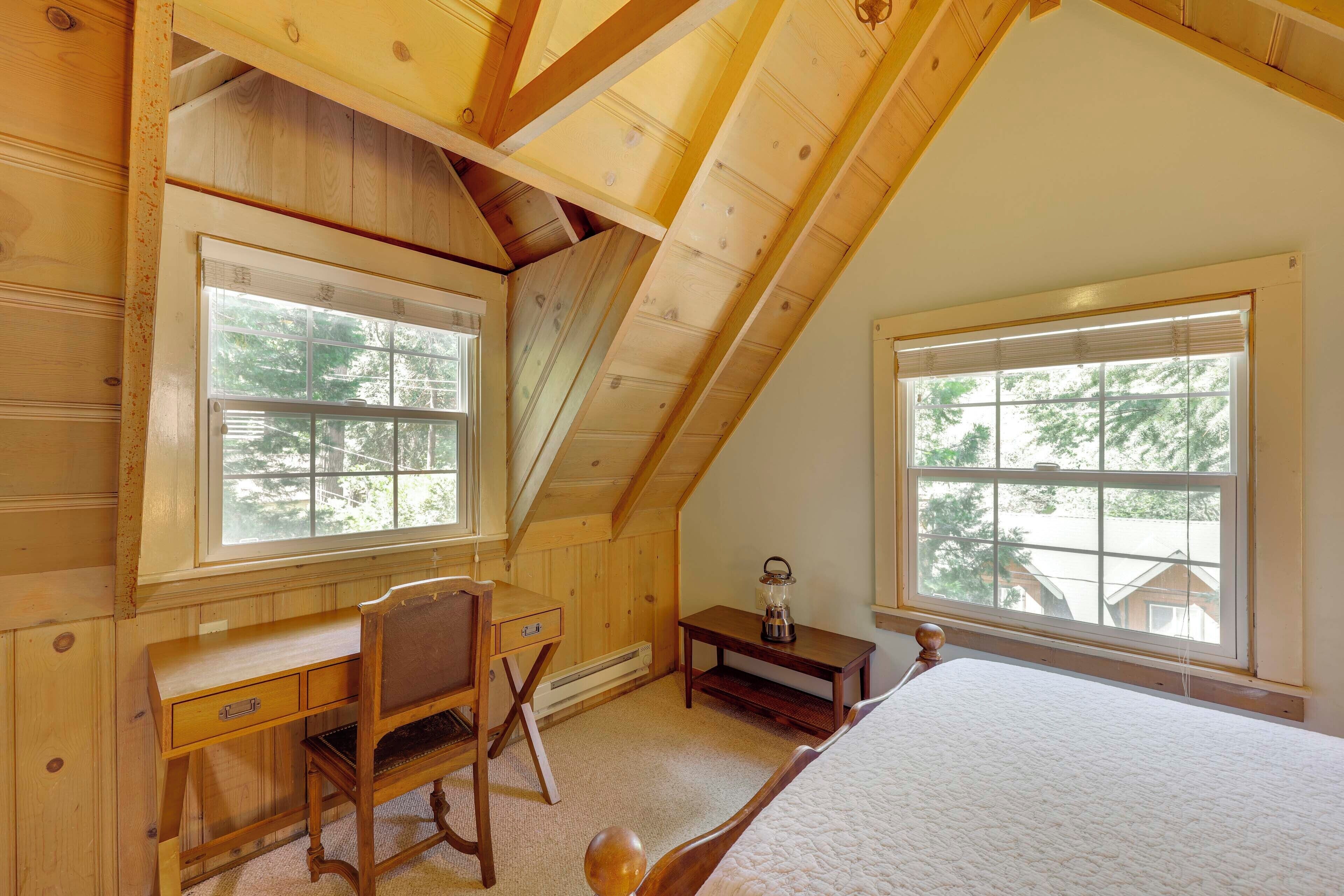 A bedroom with a sloped ceiling, wooden walls, a bed, a desk, and two windows with blinds.
