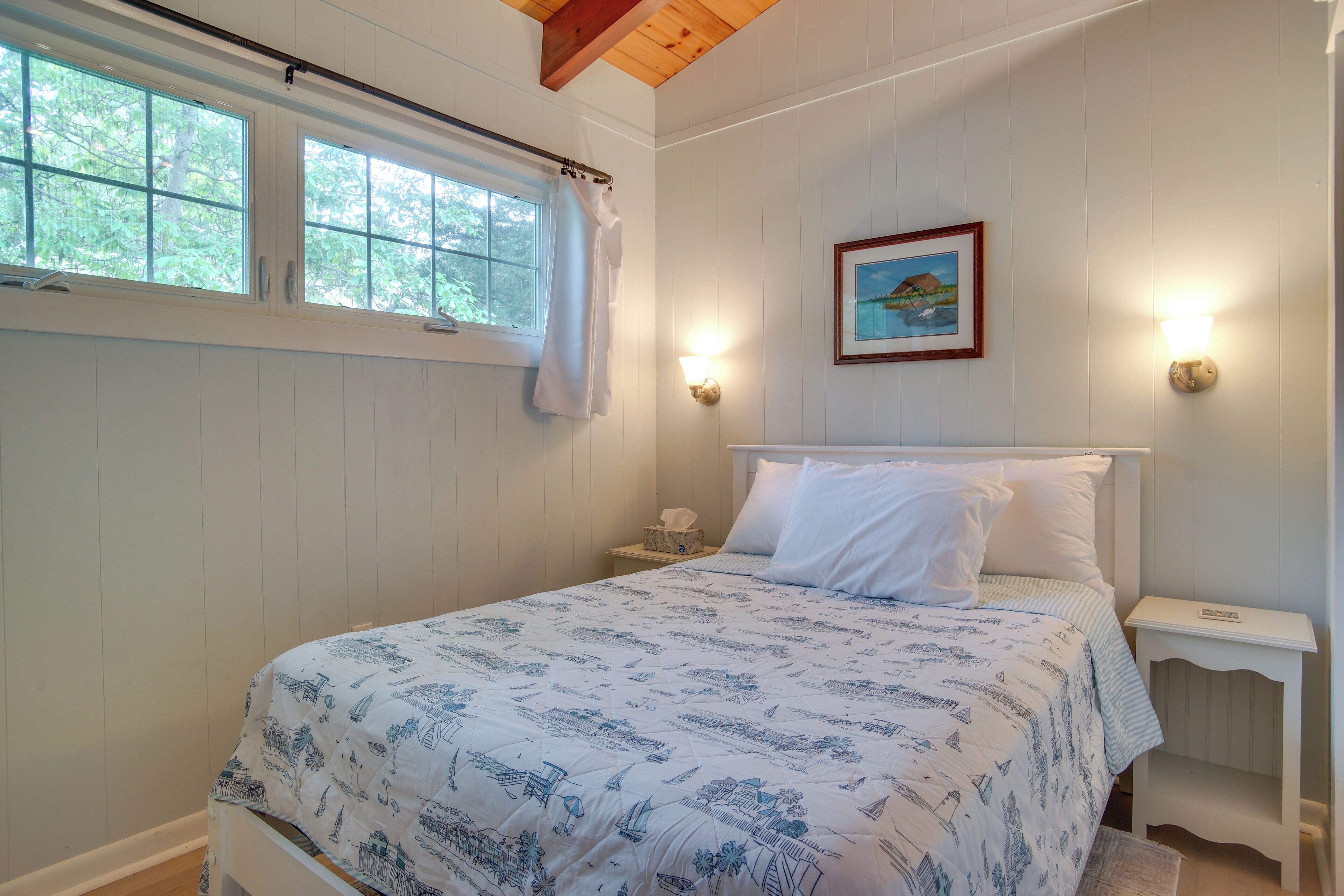 A bedroom with a bed, a nightstand, a wall-mounted light, a window with a curtain, and a framed picture on the wall.