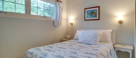 A bedroom with a bed, a nightstand, a wall-mounted light, a window with a curtain, and a framed picture on the wall.