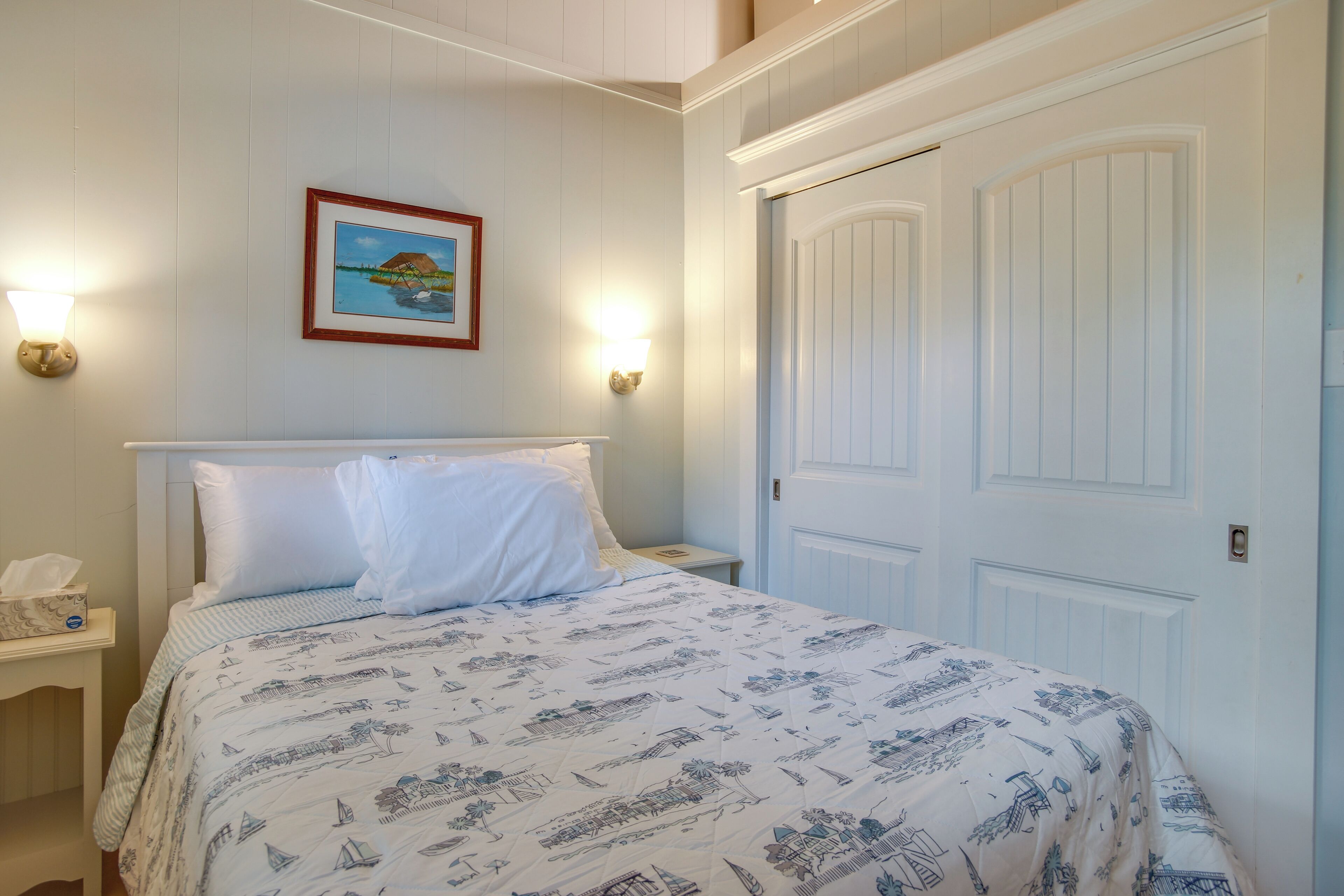 A neatly made bed with a patterned bedspread, a bedside table with a lamp, a framed picture on the wall, and a white door.
