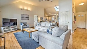 A spacious living room with a large grey sofa, a coffee table, and a TV. There is a ceiling fan, a chandelier, and a kitchen in the background. - Spacious Waterfront Falmouth Home on Jenkins Pond! (East Falmouth)