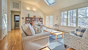 A spacious living room with a high ceiling, a skylight, a wooden floor, a sofa, a patterned armchair, a coffee table, and a bookshelf. - Spacious Waterfront Falmouth Home on Jenkins Pond! (East Falmouth)
