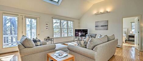 A spacious living room with two sofas, a coffee table, and a TV. Large windows provide natural light and a view of the outdoors.