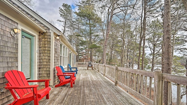 A wooden deck with two red Adirondack chairs, a wooden railing, and a view of a forested area. - Spacious Waterfront Falmouth Home on Jenkins Pond! (East Falmouth)