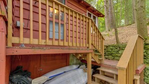 A cabin with a wooden staircase leading to a deck, a covered area with a canoe, and a stone wall in the background. - Rustic Berkshires Cottage at Lake Buel w/ Kayaks! (Great Barrington)