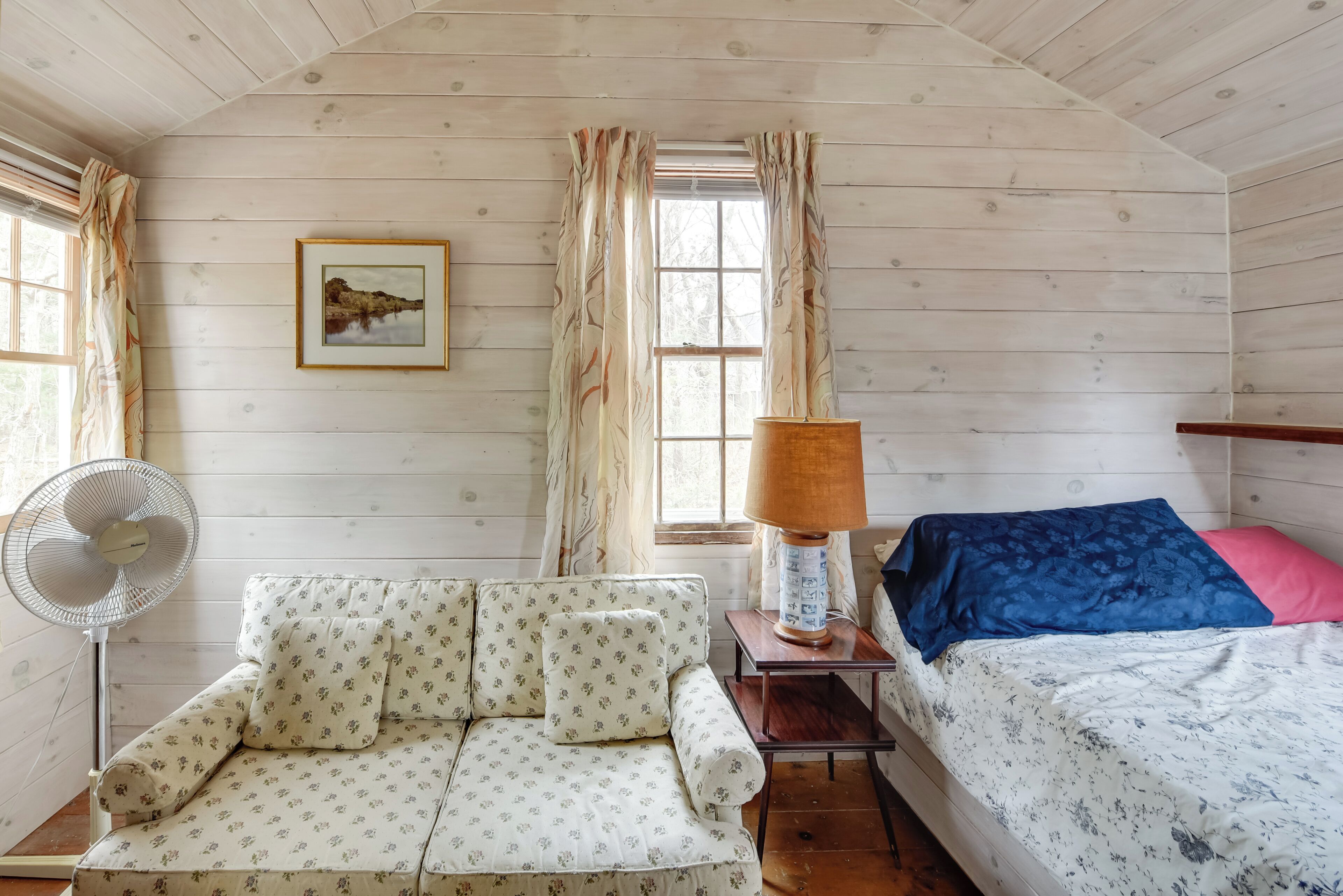 Una habitación acogedora tipo cabaña con interior de madera, un sofá, una cama con ropa de cama floral, un ventilador y una lámpara.