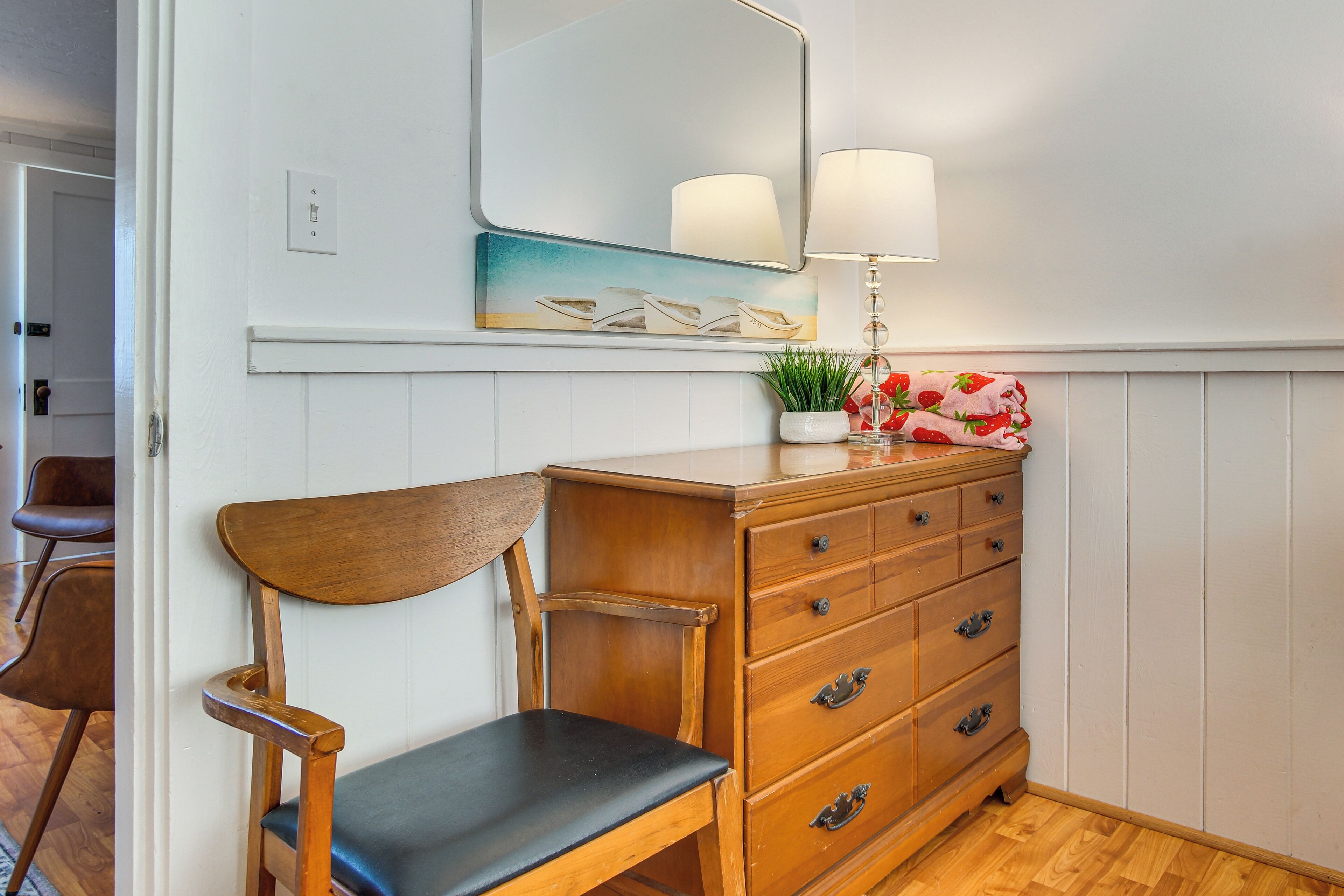 A wooden chair with a black cushion, a wooden dresser with multiple drawers, a mirror with a white frame, and a lamp with two shades.