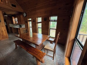 Interior de una cabaña de madera con una gran mesa de comedor, bancos y una zona de cocina.