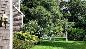 A garden with two Adirondack chairs, a lamp post, and a house with a wooden exterior. - 1 Mi to Nauset Beach: Upscale Orleans Home w/ Deck (Orleans)