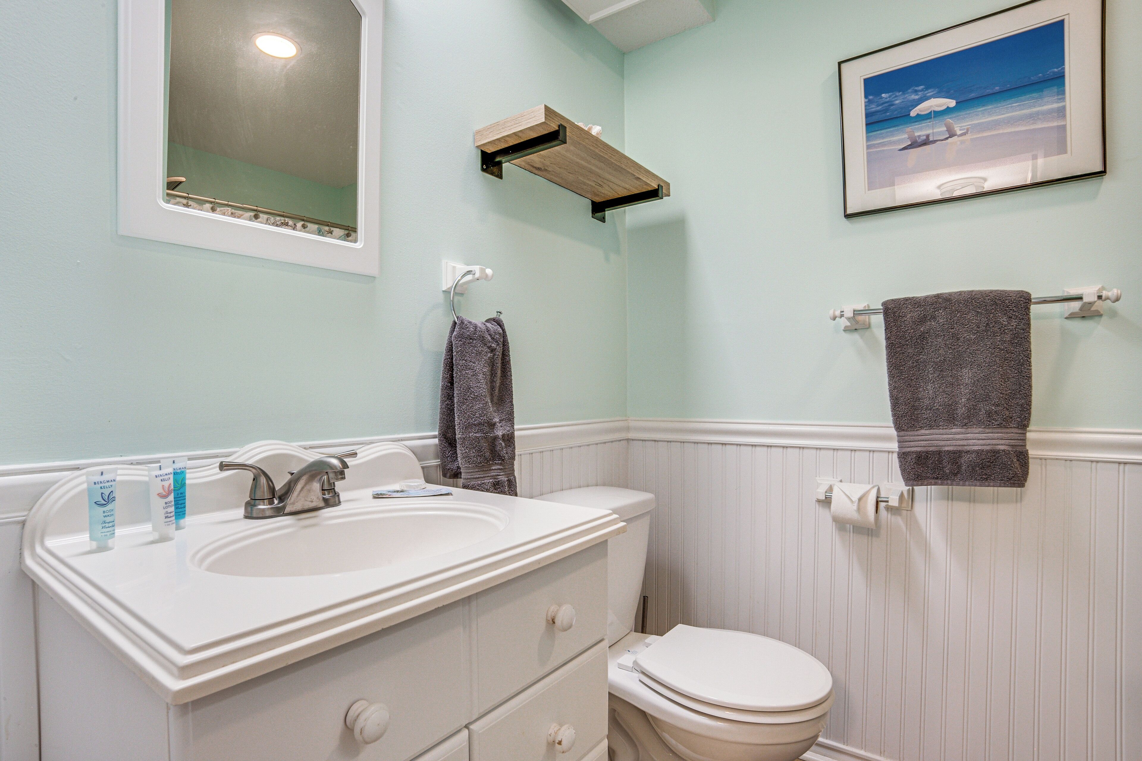 A bathroom with a white sink, mirror, and toilet, wooden shelf, and a framed picture on the wall.