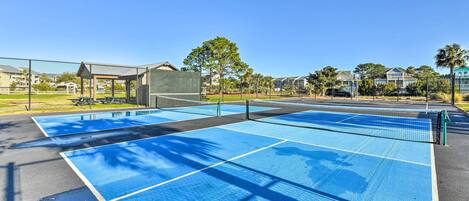 Un court de tennis avec un filet, entouré d’une clôture, et un pavillon en arrière-plan.
