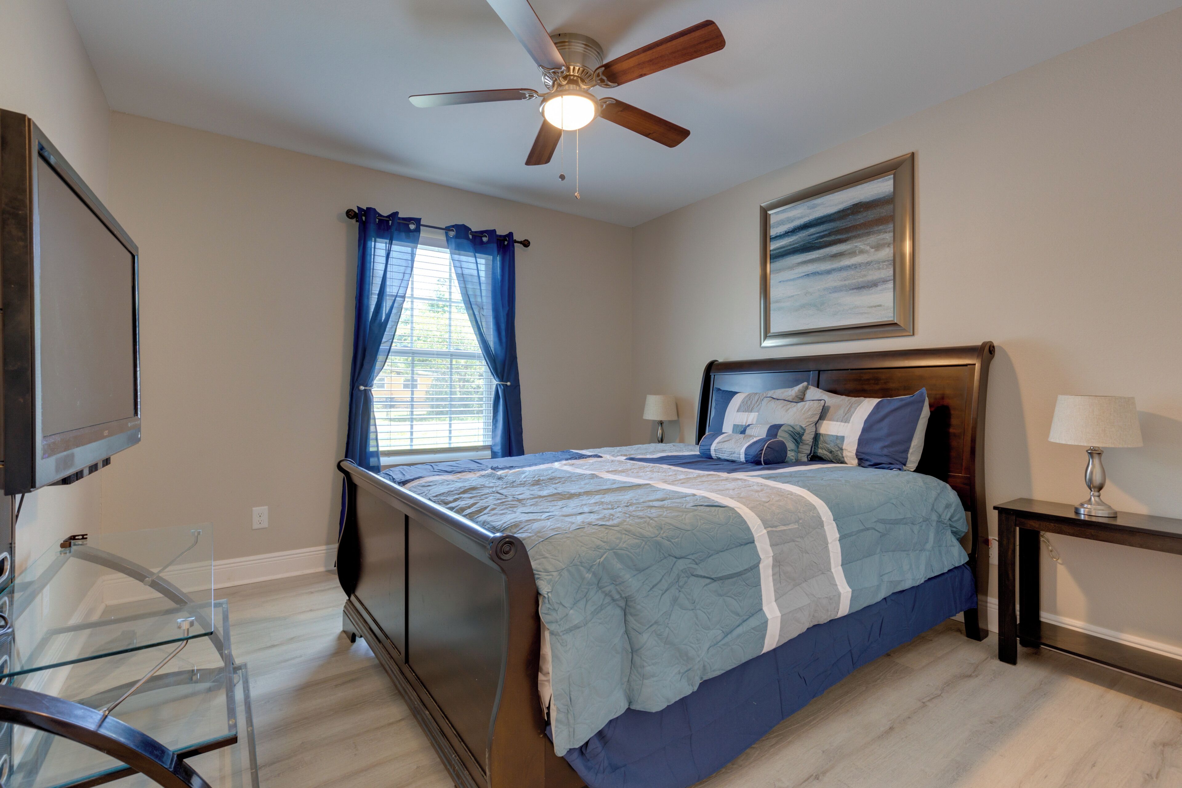 A bedroom with a bed, a ceiling fan, a window with curtains, a bedside table with a lamp, and a framed picture on the wall.