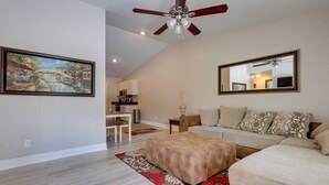 A spacious living room with a large painting, a ceiling fan, a sofa, a coffee table, and a dining area with a table and chairs. - Dog-friendly Pensacola Duplex ~ 7 Mi to Downtown (Pensacola)