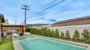 Ein rechteckiger Swimmingpool mit Fliesen umgeben, mit einer gepflegten Hecke und einer Terrasse mit Gartenmöbeln.