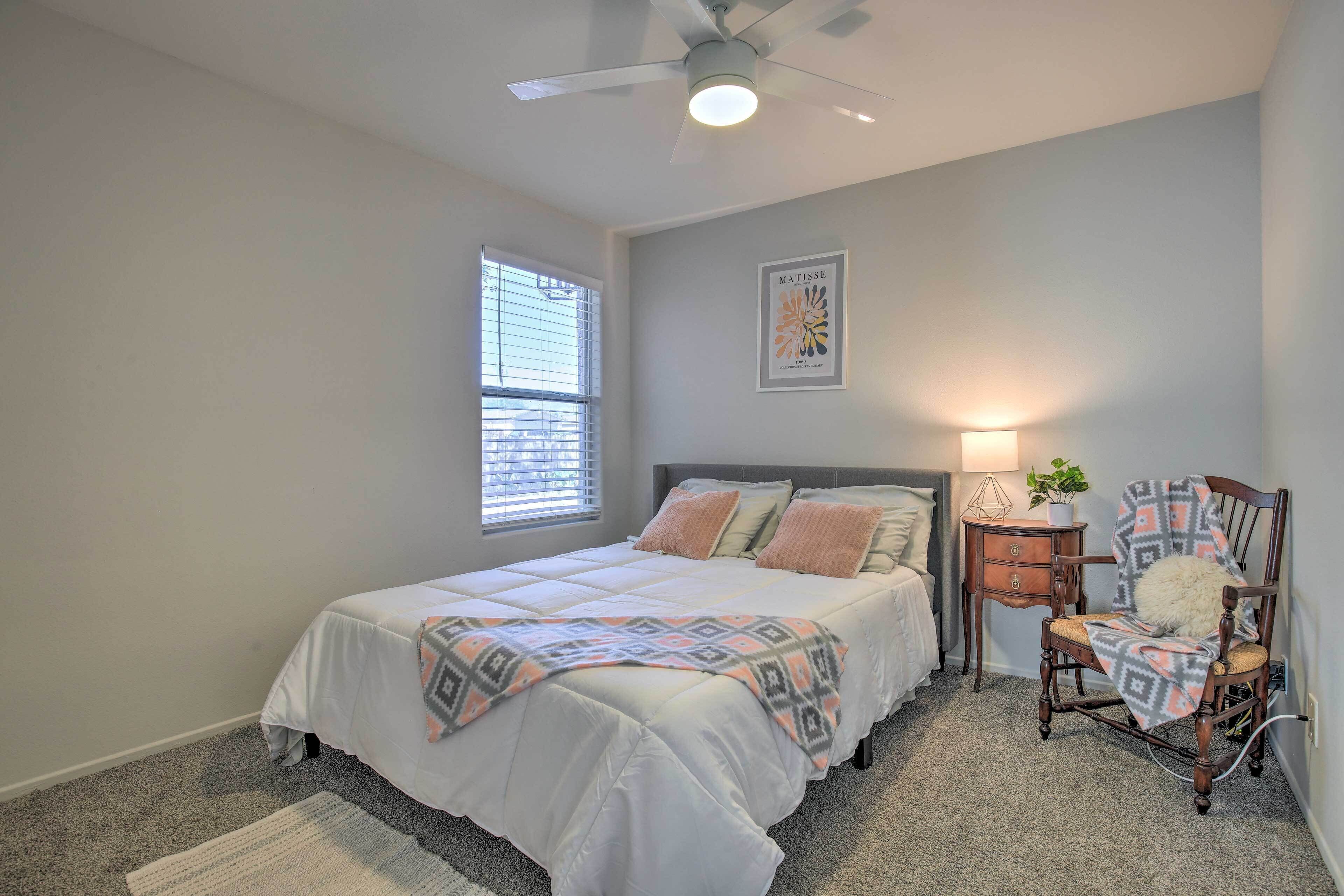 A bedroom with a bed, a chair, a nightstand, a ceiling fan, a window, and a wall art piece.