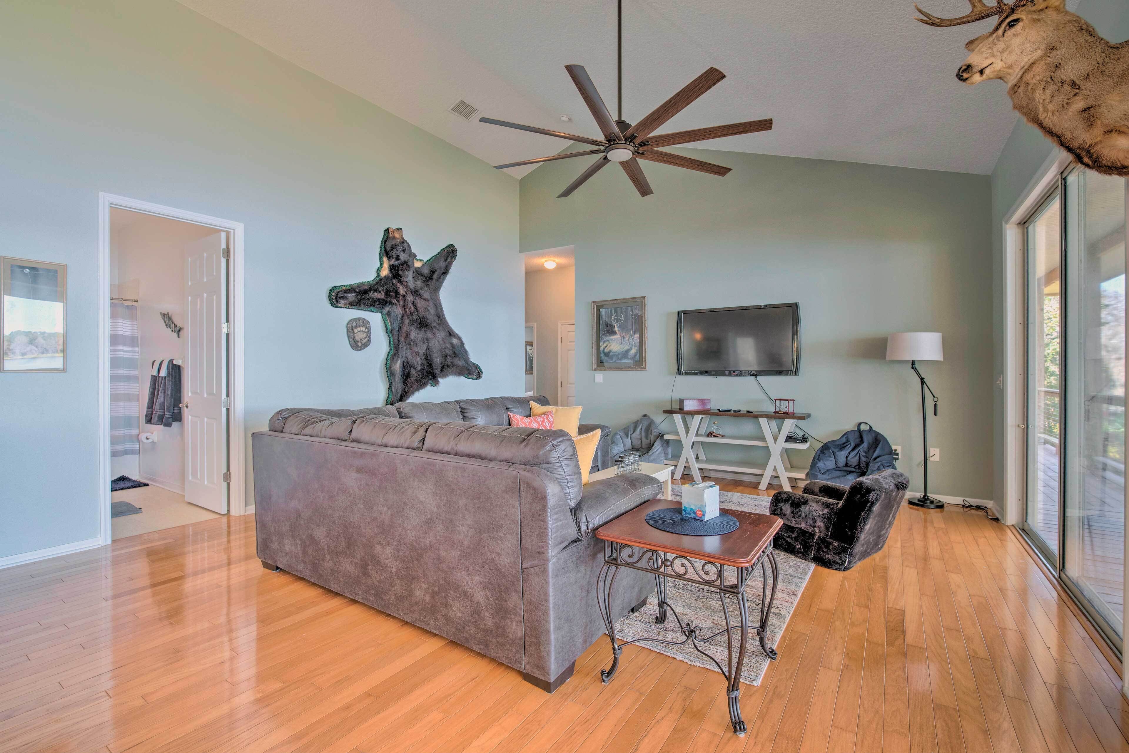 A living room with a grey sofa, a ceiling fan, a mounted deer head, a TV, and a large window.