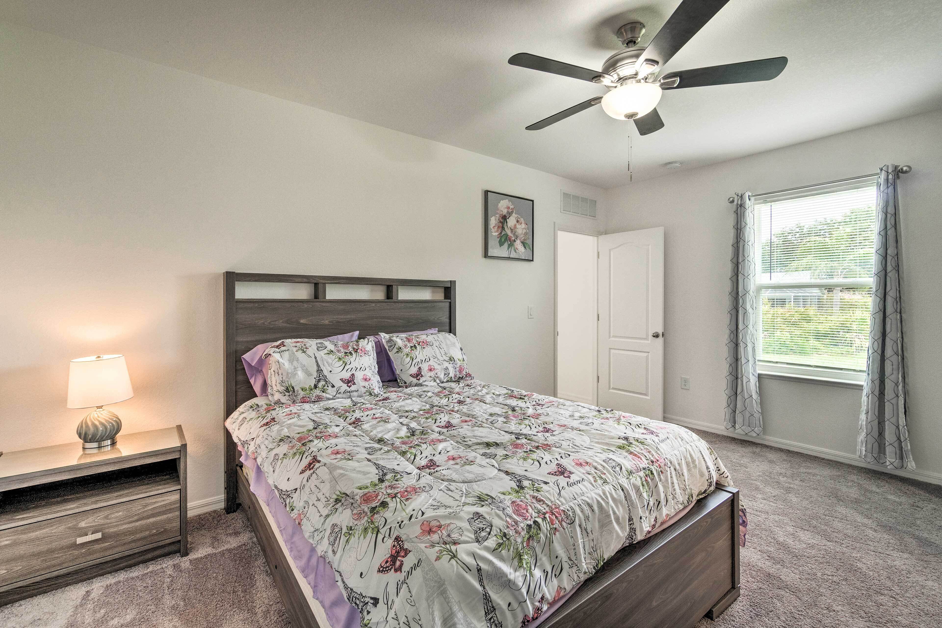 A bedroom with a bed, nightstand, lamp, ceiling fan, window with curtains, and a door.