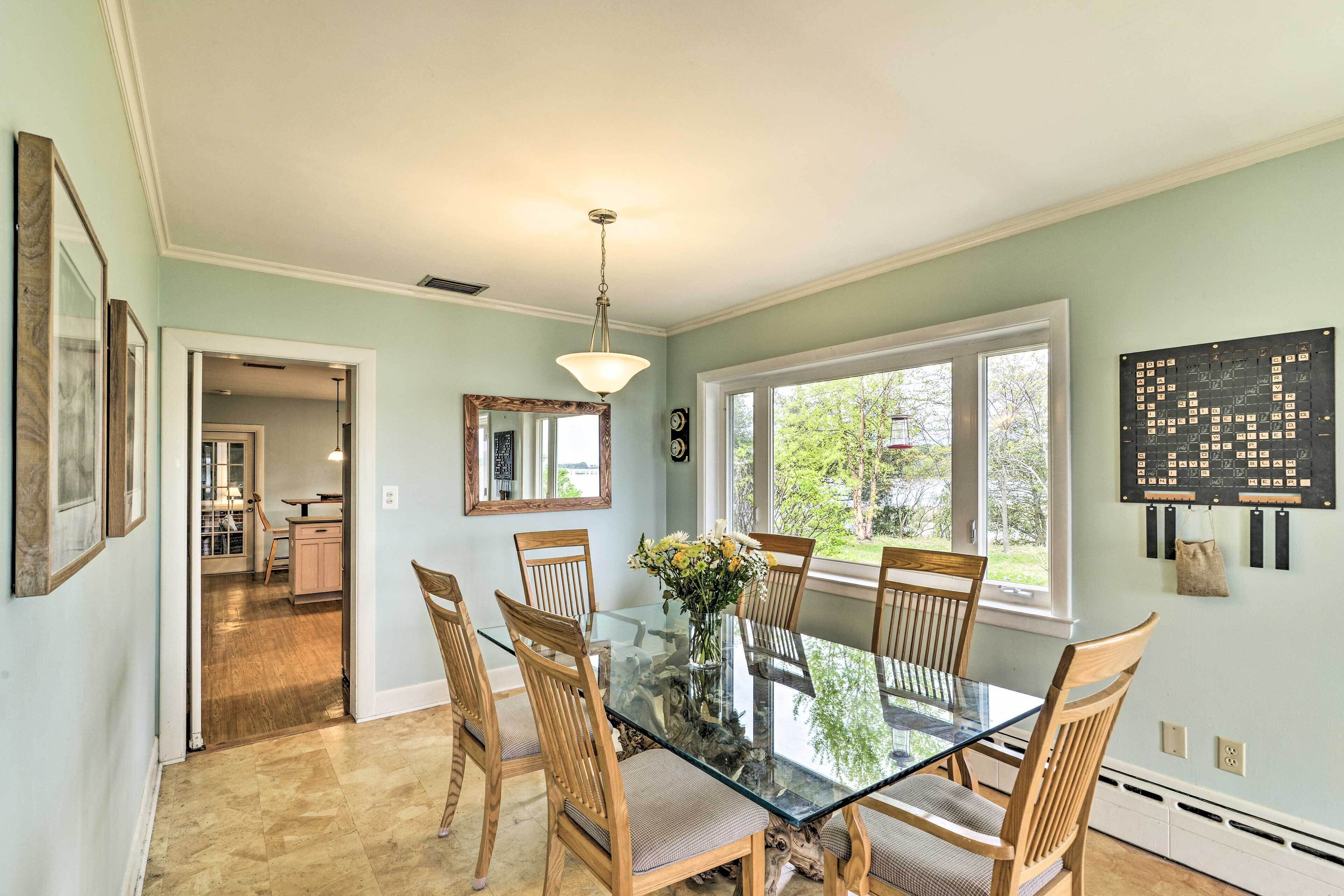 A dining room with a glass table, wooden chairs, and a vase of flowers. There is a window with a view of greenery outside.