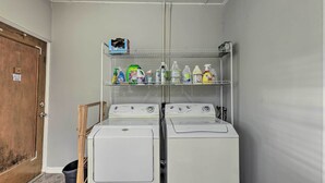 Een wasserette met twee witte wasmachines, een plank met schoonmaakmiddelen, en een houten deur.