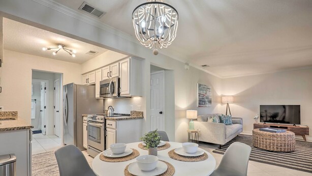 A modern kitchen with white cabinets, a microwave, and a refrigerator. There is a dining area with a white table and chairs, and a chandelier above. - Sunny St Augustine Condo w/ Wifi - Walk to Beach! (St. Augustine)