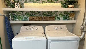 Two front-loading washing machines with control panels, a shelf above with a 'Wash Dry' sign, a blue patterned curtain, and a plant on the wall. - Sunny St Augustine Condo w/ Wifi - Walk to Beach! (St. Augustine)