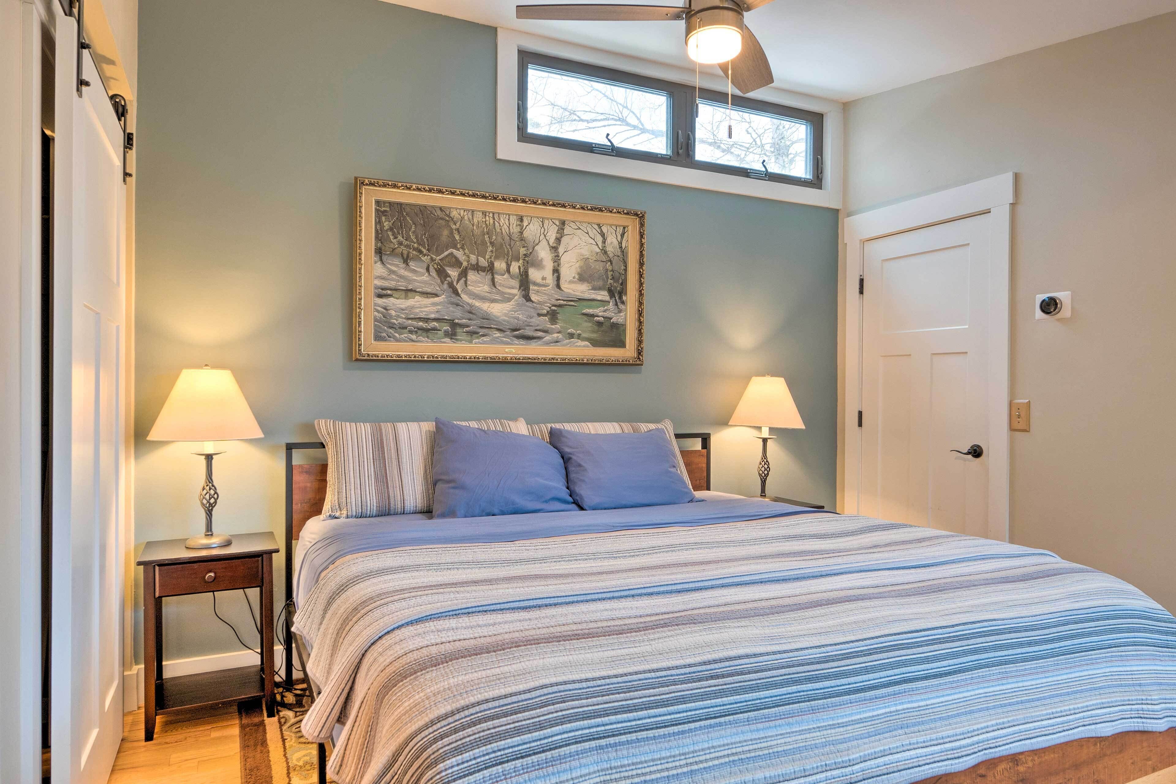 A bedroom with a bed, two bedside lamps, a ceiling fan, a window, and a door.