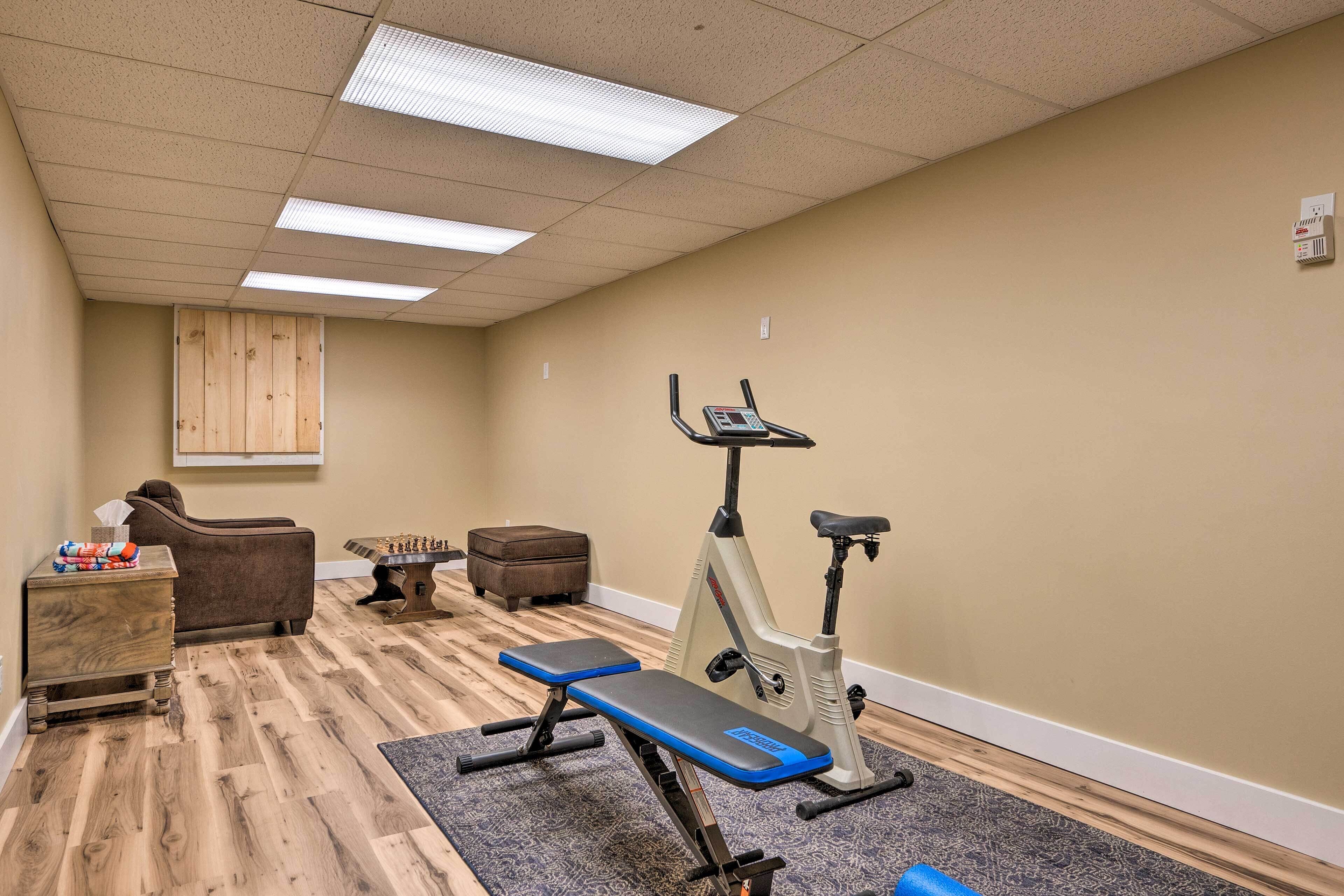 Quarto com piso de madeira, um banco, bicicleta ergométrica, cadeira de massagem e uma pequena mesa com caixa de lenços.