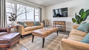 Ein Wohnzimmer mit einer Couch, einem Hocker und einem Couchtisch. Ein Fernseher ist an der Wand montiert und es gibt ein großes Fenster mit Vorhängen.