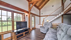 A spacious living room with a large window, wooden beams, a staircase, and a gray sectional sofa.