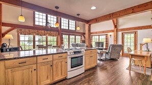 A spacious kitchen with wooden cabinets, a large island, and a built-in oven. There are pendant lights, large windows, and a dining area with a table and chairs.