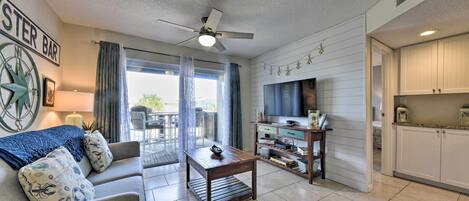A living room with a sofa, coffee table, and a TV. There is a dining area with a table and chairs. The room has a ceiling fan and a kitchen area with cabinets.