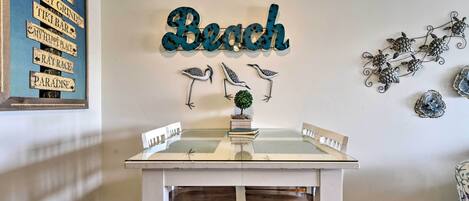 Un área de comedor con una mesa y sillas blancas, un cartel decorativo que dice "Beach" y arte de pared con pájaros y una tortuga.