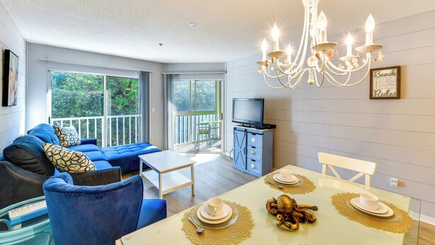 A modern living room with a blue sofa, a dining table set with white plates and gold placemats, and a chandelier. - Walk to Beach: Home w/ Porch on Anna Maria Island! (Holmes Beach)