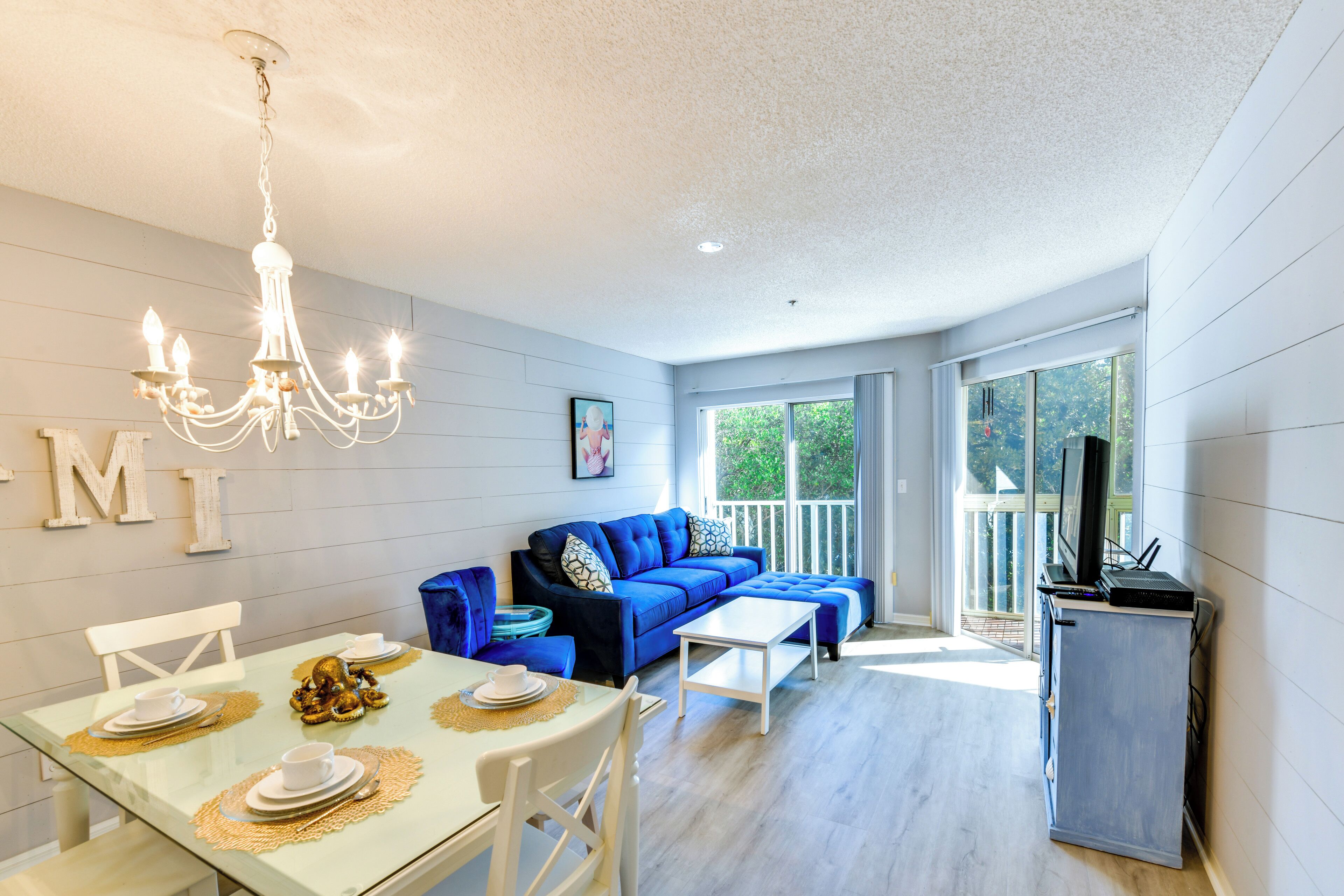 Een moderne woonkamer met een blauwe bank, een eettafel gedekt met borden en kopjes, een kroonluchter en een televisie op een standaard.